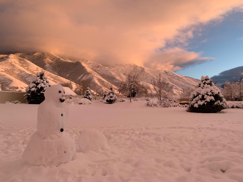 Snow scene with snowman in the foreground and snowcapped mountains in the background below sunset pink clouds